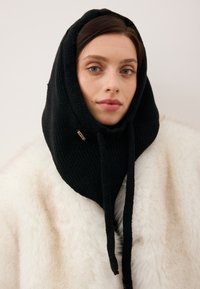 Czarna dzianinowa kaptur z troczkami i złotym akcentem. Nosi się go na białym futrzanym ubraniu z teksturowanym wzorem.