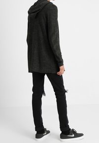 Lång, mörkgrå stickad cardigan med huva, i kombination med rivna svarta jeans och mörka sneakers, vilket visar en avslappnad lager-på-lager-look från baksidan.