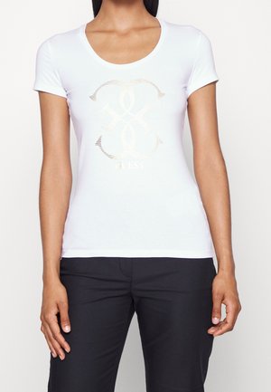 Femme portant un t-shirt blanc ajusté à manches courtes avec un logo Guess métallique doré et un pantalon noir, debout devant un fond uni.