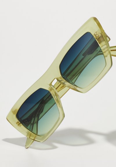 Durchsichtige gelbe Sonnenbrille mit übergroßen, winkligen Rahmen und Verlaufslinsen, die von dunklem Blau zu hellem Grün übergehen.