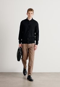 Μαύρο cardigan μακρυμάνικο με κουμπιά, σε συνδυασμό με μπεζ παντελόνι. Το μοντέλο κρατάει μια μαύρη δερμάτινη τσάντα και φοράει μαύρα παπούτσια. Απλός και μοντέρνος σχεδιασμός.