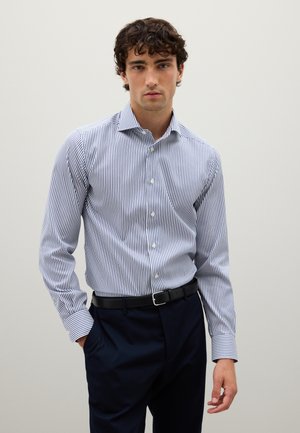 Camisa de botones a rayas en blanco y azul marino, con un cuello clásico, mangas largas y un diseño ajustado, combinada con pantalones oscuros.