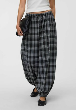 Persoon draagt losse zwart-wit geruite broek met elastische enkels, een wit topje, zwarte platte schoenen en houdt een zwarte clutch vast.