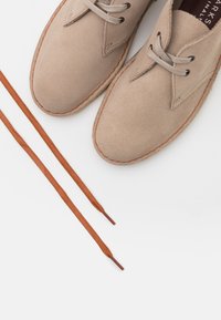 Scarpe in suede beige con cuciture marrone chiaro e lacci grigi, accompagnate da due lacci marroni disposti piatti su una superficie bianca.