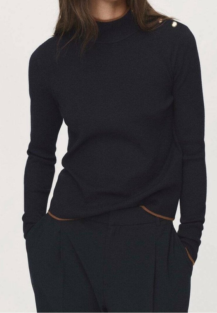 Personne portant un pull noir ajusté à manches longues avec des boutons dorés sur l'épaule et un pantalon noir taille haute, les mains dans les poches.