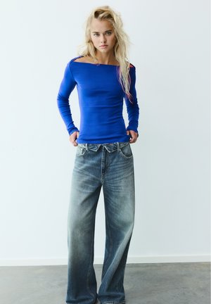 Jeune femme aux longs cheveux blonds portant un haut bleu moulant à épaules dénudées et un jean large bleu délavé, debout contre un mur blanc uni.
