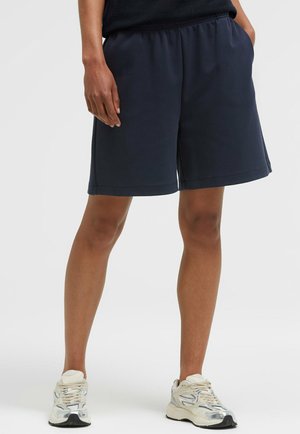Person trägt schwarze elastische Shorts und weiße Sportschuhe und steht mit den Händen in den Taschen vor einem einfarbigen Hintergrund.