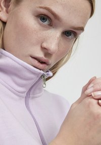 Fialová mikina na zip s vysokým límcem, texturovanou látkou a stříbrným detailem na zipu. Detailní záběr na ruce, které se dotýkají.