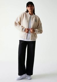 Kvinna iklädd beige fleecejacka med New York Yankees-logotyp, vit skjorta, svarta vida byxor, vita sneakers och svart baseballkeps.