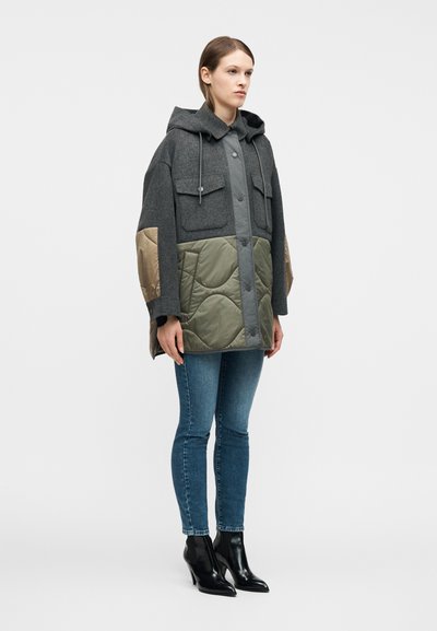 Γυναίκα που φοράει παλτό με κουκούλα και χρωματικό μπλοκ, με επένδυση σε χακί κατω μέρος και γκρι μαλλί στο πάνω μέρος, συνδυασμένο με μπλε τζιν και μαύρα μποτάκια αστραγάλου.