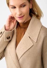 Beige wollen mengsel jas met grote revers en bruine knopen, gedragen over een bruine geplooide top. De jas heeft een zachte, gestructureerde afwerking.
