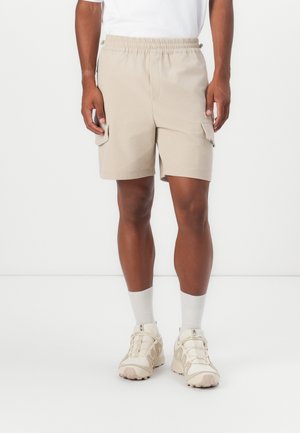 Beige cargo shorts with an elastic waistband, side pockets, and a smooth fabric texture. Paired with white socks and light-colored sneakers.