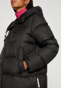 Veste matelassée noire avec capuche, dotée d'une fermeture éclair frontale, de cordons resserrés et d'une poche latérale avec une étiquette logo. Texture lisse et brillante.