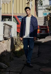 Sötétkék farmering, alatta fehér póló, kék farmernadrággal és fekete cipővel párosítva. A háttérben egy piros autó és egy fehér kerítés látható.