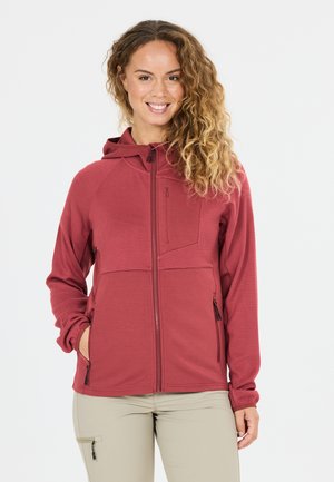 Lächelnde Frau mit lockigem Haar, die eine rote, strukturierte Kapuzenjacke mit Reißverschluss und beige Hosen mit Reißverschlusstaschen trägt.