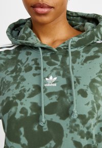 Grön hoodie med ett tie-dye-mönster, med en vit Adidas-logga på bröstet och vita ränder på ärmarna. Mjuk tygtextur.