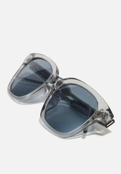 Ochelari de soare din plastic transparent cu lentile de culoare albastră închisă. Design unghiular, ramă groasă și accesorii subtile din argint pe brațe.