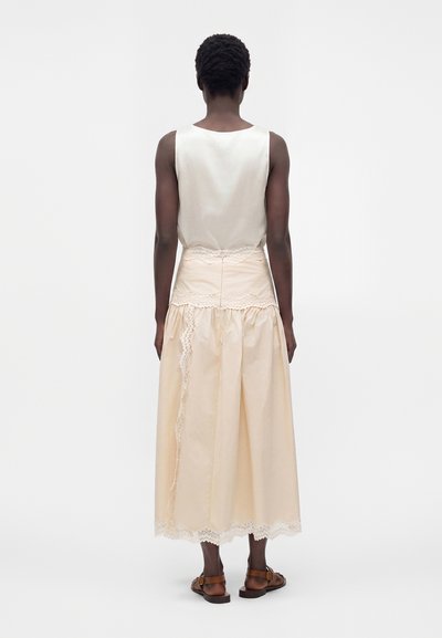 Femme aux cheveux courts et bouclés, portant un haut blanc sans manches et une jupe midi beige bordée de dentelle, debout de dos, sandales marron visibles.