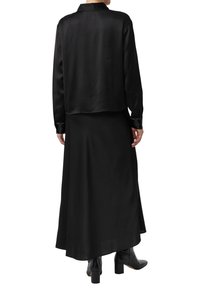 Person wearing a black long-sleeve satin blouse and matching black maxi skirt with black heeled ankle boots, seen from the back.