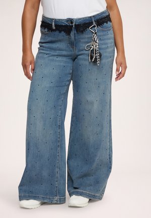 Persona che indossa jeans blu a gamba larga con piccoli pois neri, bordo in pizzo nero in vita, portachiavi decorativo, scarpe bianche e top bianco.