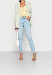 Persona che indossa un blazer verde chiaro, un top arricciato floreale giallo, jeans strappati blu chiaro, tacchi aperti arancioni e tiene in mano una borsa bianca.