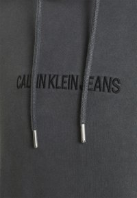 Sudadera gris con el logotipo bordado en negro "CALVIN KLEIN JEANS". Incluye cordones texturizados con puntas metálicas y una textura de tela suave.