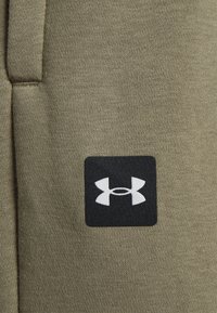 Olivgrüner Stoff zeigt eine Nahaufnahme eines schwarzen quadratischen Aufnähers mit einem weißen Logo. Die Textur ist glatt und leicht dehnbar.