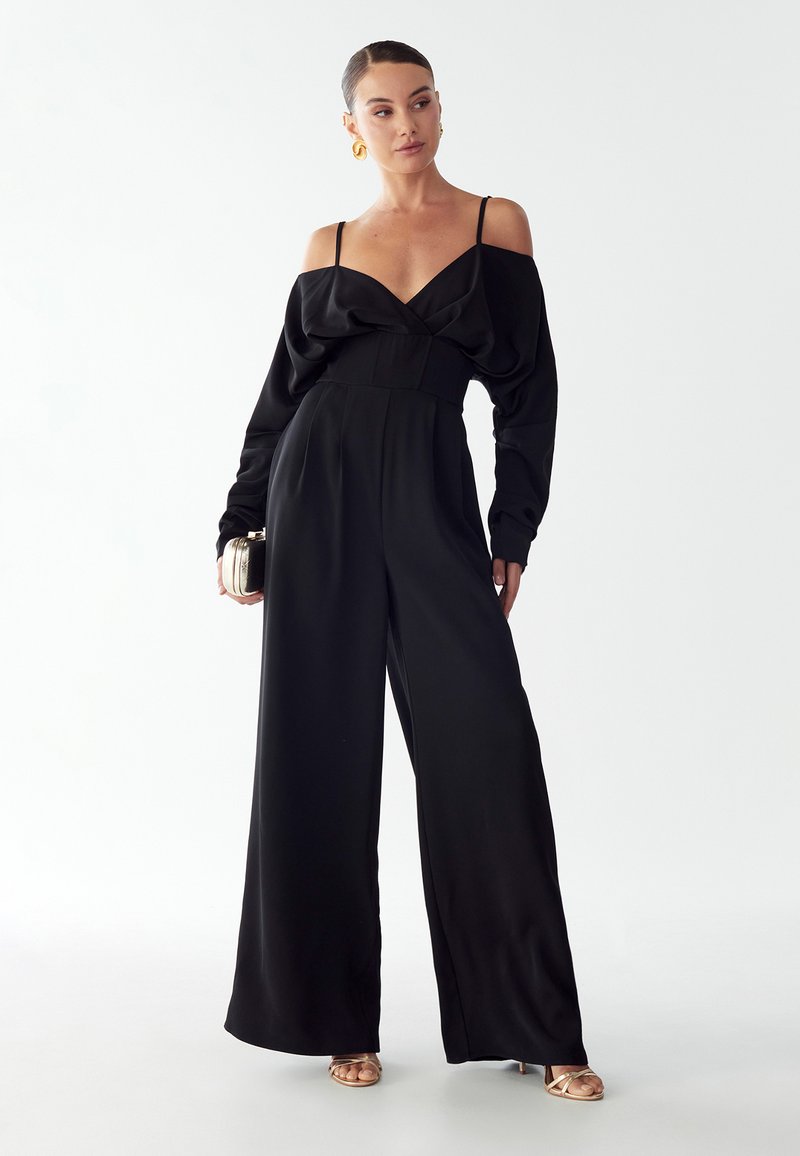 Moteris dėvi juodą šalia pečių sukurtą šokinėjimą su plačiomis kojomis, auksinius auskarus, laiko mažą metalinę rankinę ir auksinius atvirus batus ant kulno.