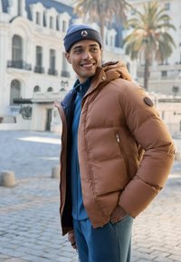 Giacca imbottita marrone con cappuccio, tasche con cerniera e fodera interna a contrasto, abbinata a una felpa blu e pantaloni, indossata all'aperto.