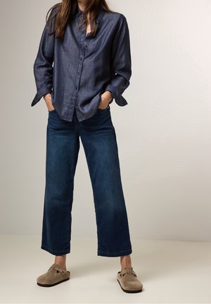 Personne portant une chemise bleu foncé à manches longues boutonnée, un jean bleu foncé à jambes larges et des chaussures marron clair sans lacets, debout les mains dans les poches.