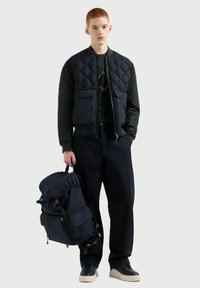 Gilet trapuntato blu navy con chiusura a zip, maniche lunghe e tasche frontali, abbinato a pantaloni scuri e uno zaino coordinato. Tessuto testurizzato.