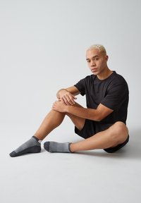 Schwarzes, kurzärmeliges Sportshirt, schwarze Shorts, graue gemusterte Socken, sitzend auf einem hellen Hintergrund, mit kurzem blonden Haar.