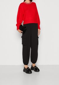 Maglione rosso lavorato a maglia con colletto di perline, abbinato a pantaloni cargo neri larghi con tasche laterali e mocassini neri lucidi.