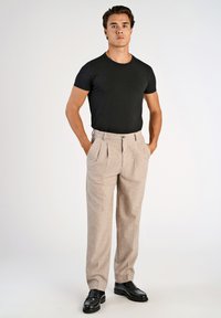 Camiseta negra ajustada y pantalones beige de pliegues hechos de un tejido texturizado, combinados con zapatos negros con cordones, de pie contra un fondo liso.