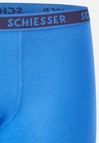 Boxers en coton bleu avec une texture côtelée et une ceinture sombre arborant le nom de la marque "Schiesser" en lettres bleu clair contrastantes.