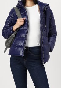 Chaqueta de plumas azul marino con un acabado brillante, cierre con cremallera y capucha, usada sobre una blusa blanca y combinada con jeans de mezclilla oscura.
