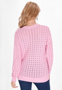 Rosa stickad tröja med ett strukturerat öppet mönster, ribbade ärmslut och nederkant. Baksidan är löst sittande med rund halsringning.