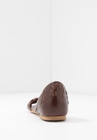 Chaussure plate en cuir marron avec un motif tressé au niveau du talon, design à bout pointu, et une semelle plate de couleur claire sur fond blanc.