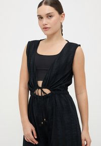 Musta jumpsuit lyhyillä hihoilla, jossa on nyörivyötärö ja leikattu yksityiskohta edessä. Rakeinen kangas, jossa on hienovaraisia raitoja.