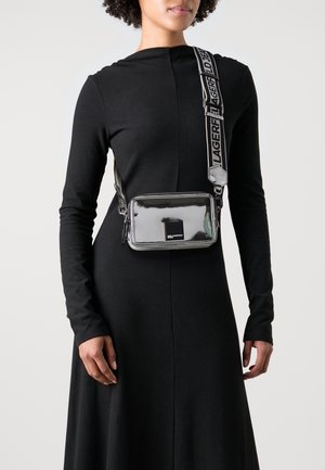 Sølv crossbody-taske med blank finish, rektangulær form og sort logo-mærkat. Har justerbar rem med kontrastfarvet tekst.