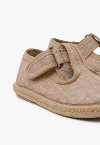 Beige fabric shoes with a Velcro strap, textured upper, and woven jute-like sole. Rounded design with contrasting stitching details.