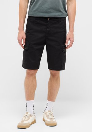 Mann trägt schwarze Cargo-Shorts, weiße Sportsocken mit Schriftzug, beige-weiße Sneakers und ein grünes T-Shirt und steht vor einfachem Hintergrund.