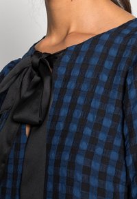 Blouse à manches courtes en bleu foncé avec un motif à carreaux et un nœud en satin noir au niveau du col. Détail en tissu texturé visible.