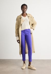 Beige trenchcoat met een relaxte pasvorm, gecombineerd met een fluffige witte trui en strakke paarse broeken. Lichtgekleurde sneakers maken de outfit compleet.