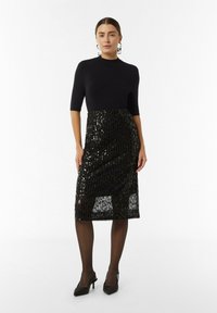 Schwarzes Kleid mit tailliertem Oberteil, kurzen Ärmeln und einem glitzernden Midi-Rock mit vertikalen Mustern. Getragen mit transparenten schwarzen Strumpfhosen und High Heels.