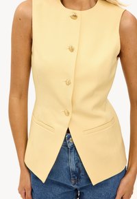 Geel mouwloos vest met een aansluitend ontwerp, voorzien van vier gouden schelpvormige knopen en twee voorzakken. Draag het met hoge taille spijkerbroekshorts.