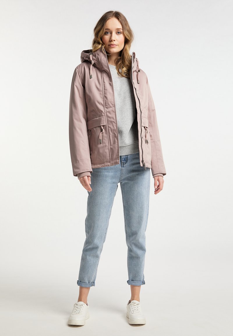 Junge Frau steht in einer hellrosa Kapuzenjacke, grauem Sweatshirt, hochgekrempelten hellblauen Jeans und weißen Turnschuhen vor weißem Hintergrund.