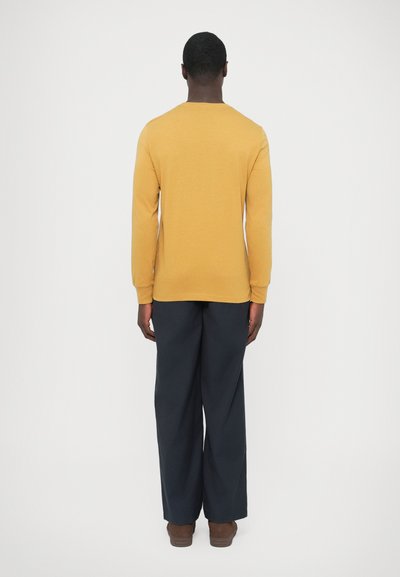 Cămașă galben-muștar cu mâneci lungi și decolteu rotund, asortată cu pantaloni largi bleumarin și pantofi maro, văzută din spate.