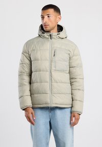 Chaqueta de plumón gris claro con capucha, costuras verticales, bolsillo en el pecho con cremallera y puños elásticos. Lleva puesta sobre unos jeans azules.