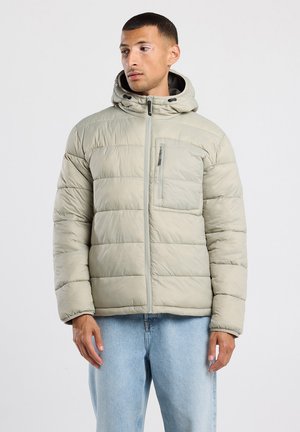 Lichtgrijze pufferjack met capuchon, verticale stiksels, ritszak op de borst en elastische manchetten. Draagt over blauwe jeans.